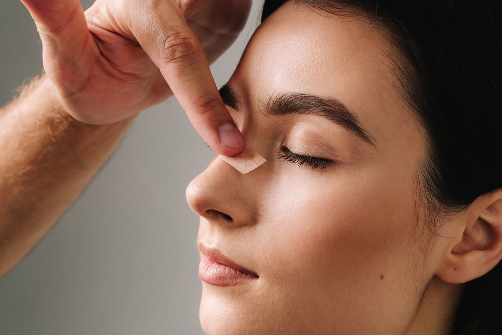 Surgeon inspecting nose tip bandage on woman’s face for potential post-rhinoplasty follow-up indicators.