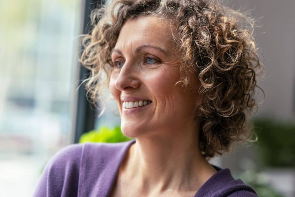 Middle-aged woman smiling near window, showing smooth skin and positive outcome from facelift recovery care.