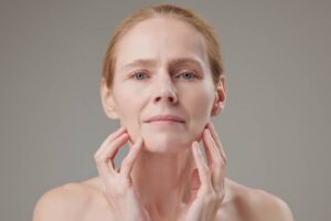 A mature woman with blonde hair looking directly at the camera while gently touching her jawline and cheeks with both hands.