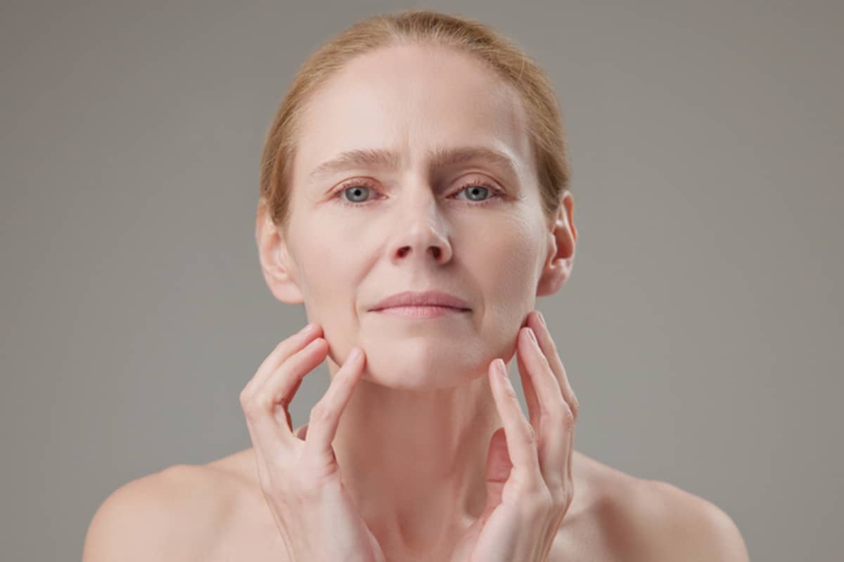 A mature woman with blonde hair looking directly at the camera while gently touching her jawline and cheeks with both hands.