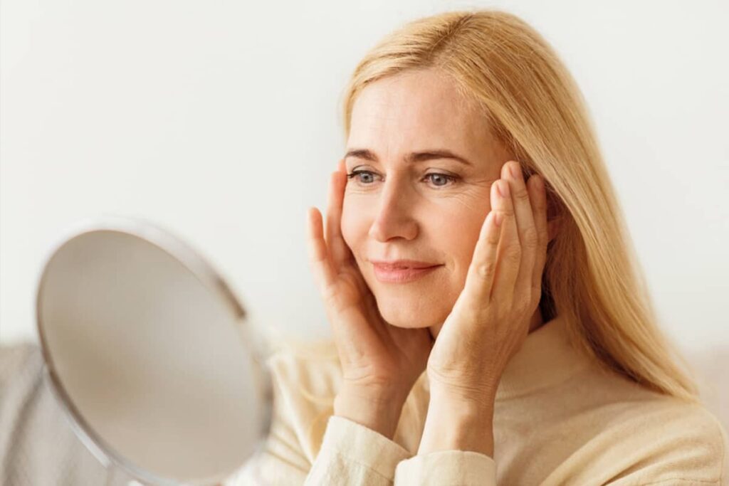 A blonde woman looking into a handheld mirror and gently lifting the skin on her temples to visualize the effects of a facelift.