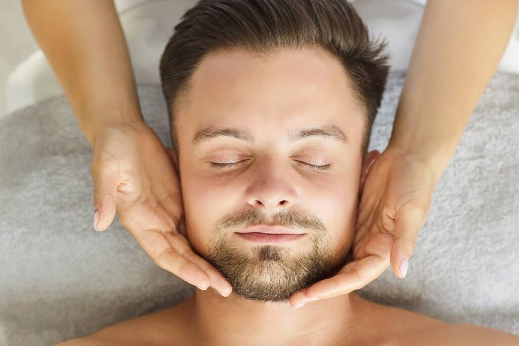 An overhead view of a relaxed man with his eyes closed receiving a therapeutic facial massage or gentle jawline assessment from a practitioner's hands.