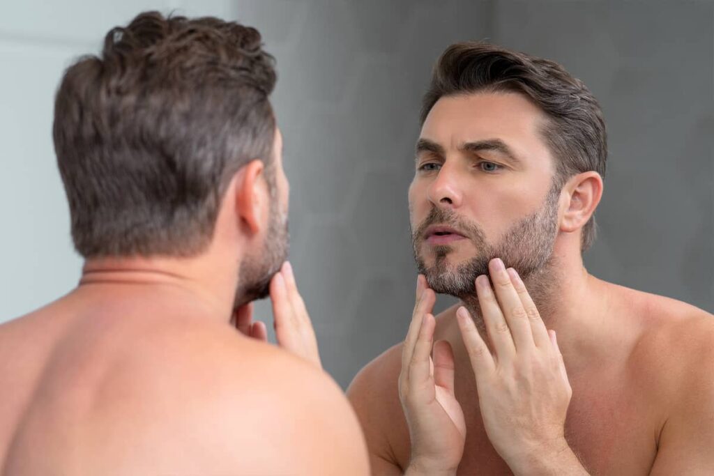 A middle-aged man with a groomed beard looking at his reflection in a bathroom mirror while gently touching his jawline and neck area.