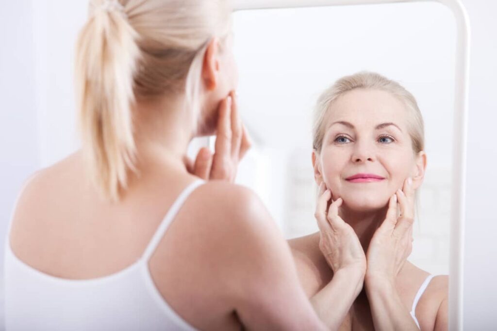 A blonde middle-aged woman looking at her reflection in a white-framed mirror while gently lifting the skin on her cheeks and jawline with both hands.