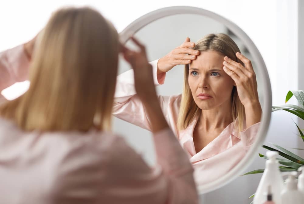 A blonde woman with medium-length hair looks into a round mirror with a concerned expression, using her fingertips to examine lines on her forehead.